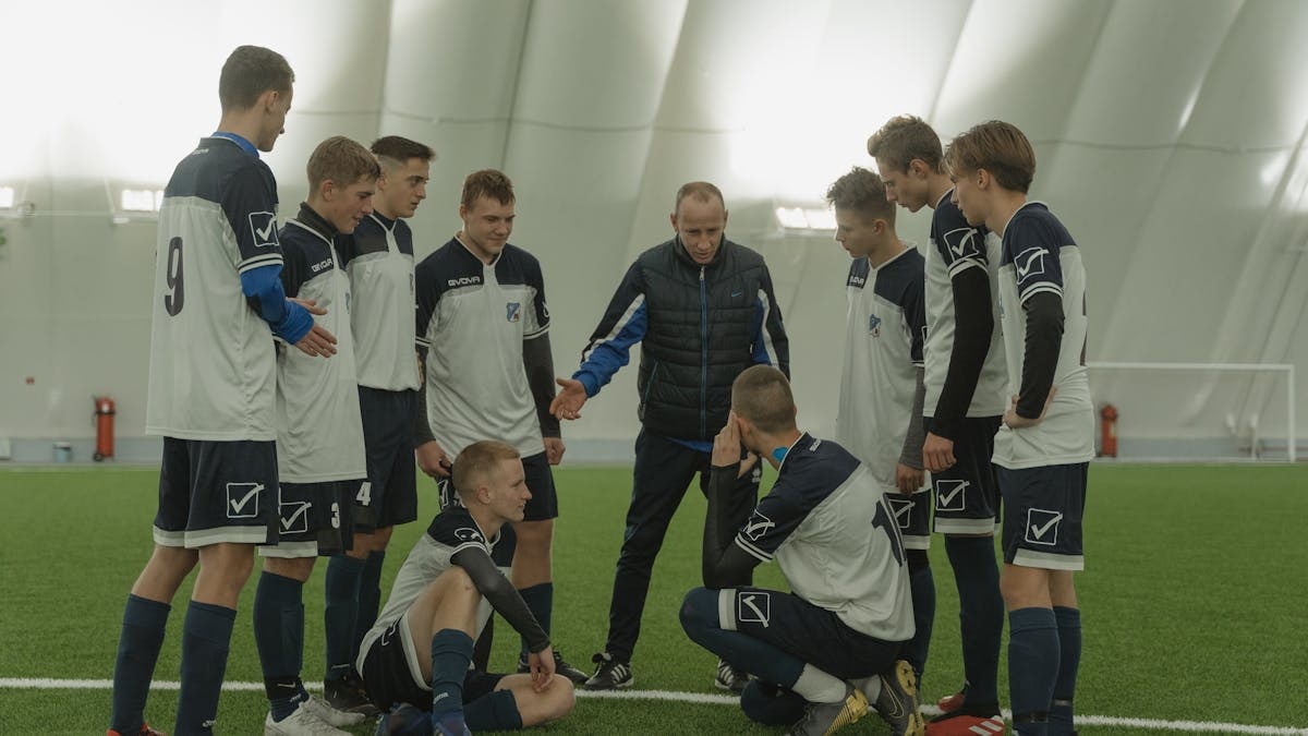 A soccer coach gives tactical advice to a youth team indoors on artificial turf.